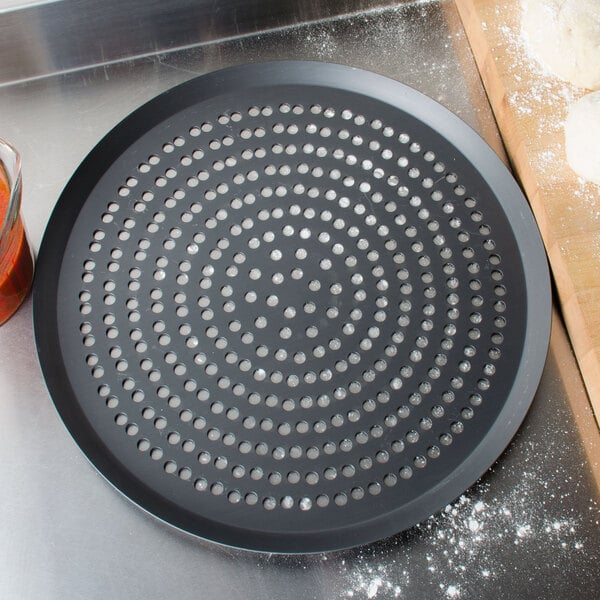 An American Metalcraft Super Perforated Hard Coat Anodized Aluminum Cutter Pizza Pan on a brown surface next to a cutting board.