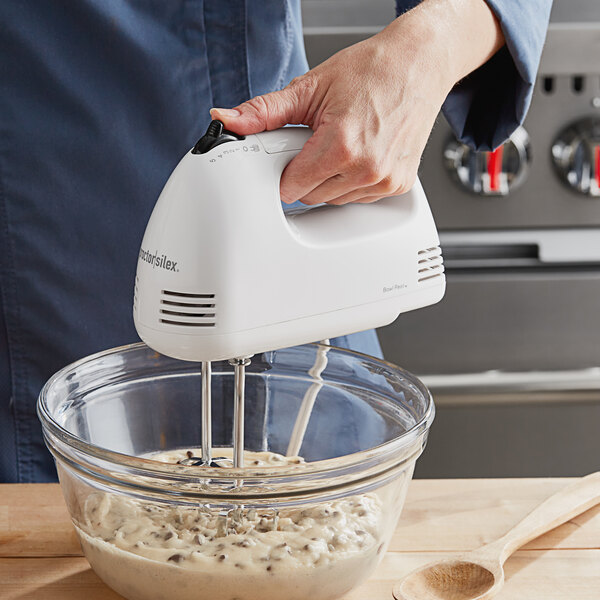 A person using a white Proctor Silex hand mixer to mix white batter in a bowl.