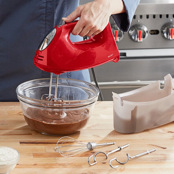 A person using a red Hamilton Beach hand mixer to whisk brown liquid in a bowl.