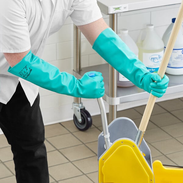 A man wearing Ansell AlphaTec green dishwashing gloves is using a mop to clean a floor.