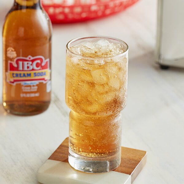 A glass of IBC Cream Soda with ice on a wooden coaster next to a bottle.