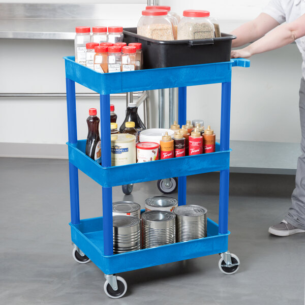 A blue Luxor plastic utility cart with three shelves holding clear containers with red lids and a bottle of liquid with a yellow cap.
