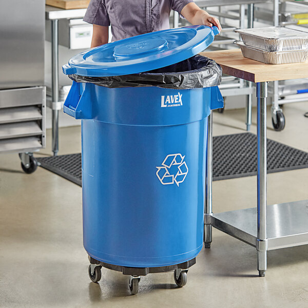 A woman standing in a school kitchen next to a Lavex blue recycling can with a blue lid.