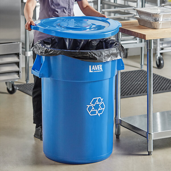 A woman in a blue uniform standing next to a Lavex blue recycling can with a blue lid.