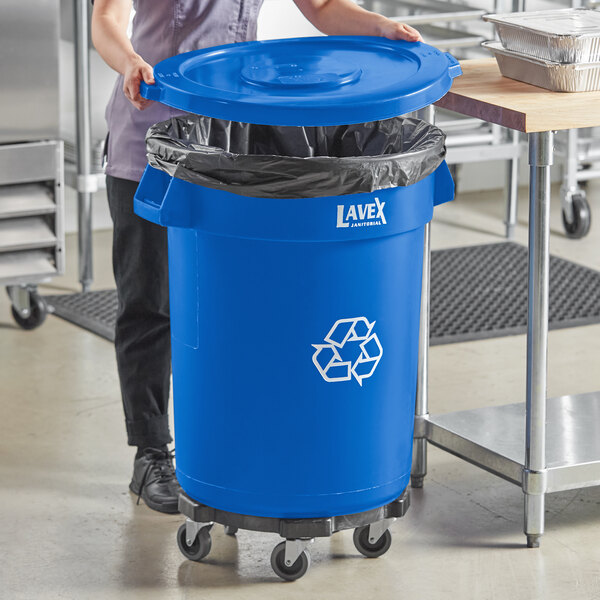 A woman holding a Lavex blue round commercial recycling can with a blue lid in a kitchen.