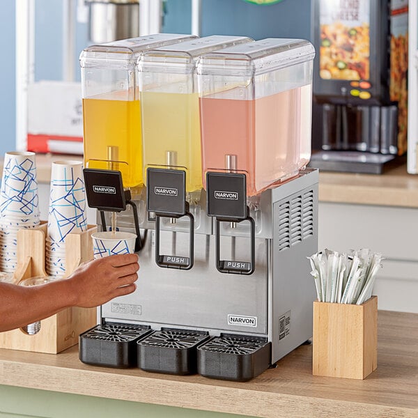 A person pouring yellow liquid into a Narvon refrigerated beverage dispenser with two other containers of colored liquid nearby.