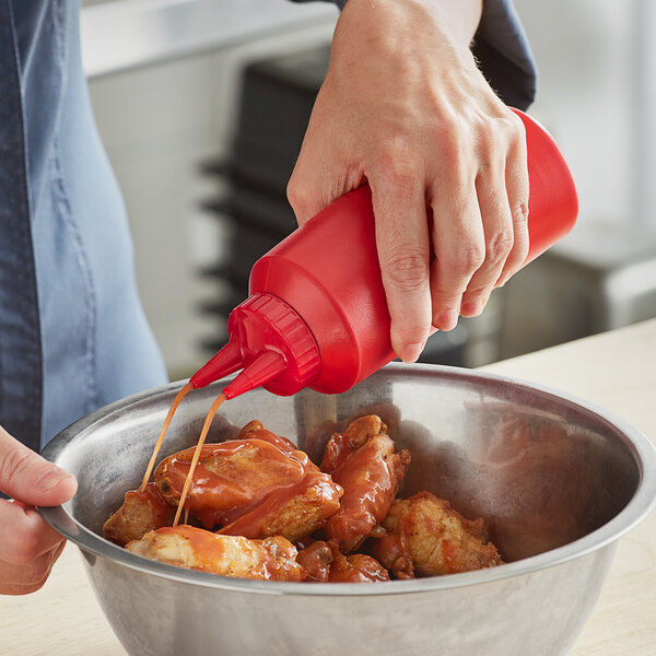 A hand holding a red Vollrath Twin Tip squeeze bottle pouring sauce into a bowl of chicken.