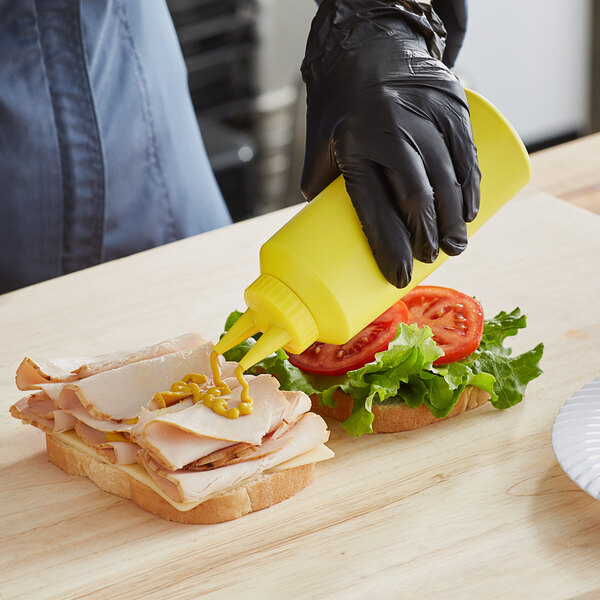 A person in black gloves using a Vollrath yellow squeeze bottle to put mustard on a sandwich with tomato, lettuce, and meat.