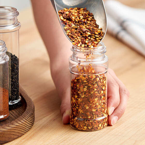 A person pouring red pepper flakes into a round plastic spice jar.