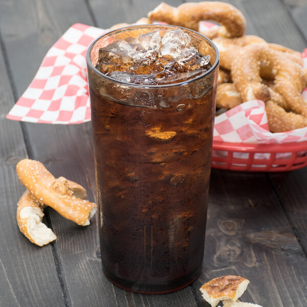 A light amber Cambro plastic tumbler filled with brown soda on a table with a basket of pretzels.