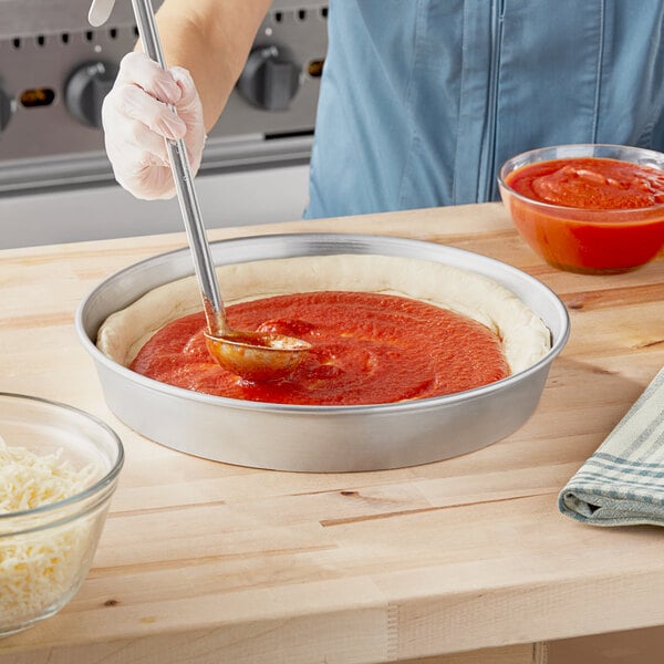 A person using a metal rod to spread sauce in a Choice aluminum tapered deep dish pizza pan.