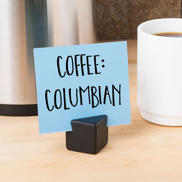 A black American Metalcraft cube card holder with a sign in it on a white surface next to a cup of coffee.