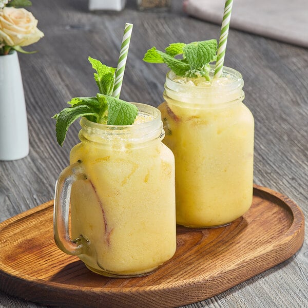 A glass jar filled with yellow Fanale peach syrup on a table with two glasses of yellow smoothie and mint leaves.