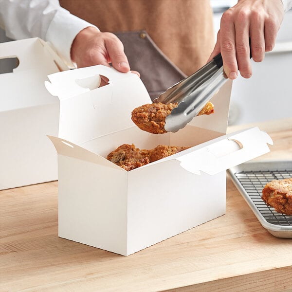 A hand holding a piece of fried chicken in a white barn take-out box.