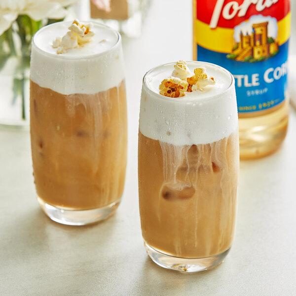 Two glasses of iced coffee with brown liquid and white foam on top, with Torani Kettle Corn Flavoring Syrup on the side.