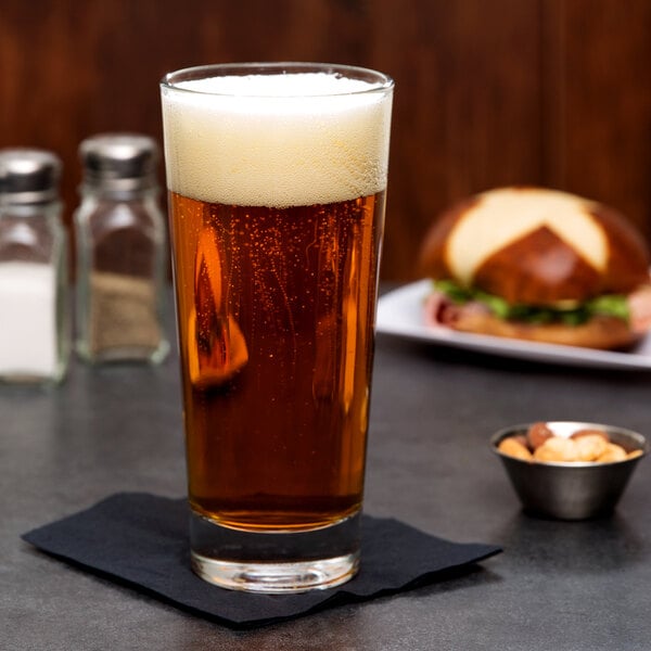 A Libbey customizable cooler glass of beer on a napkin next to a sandwich.
