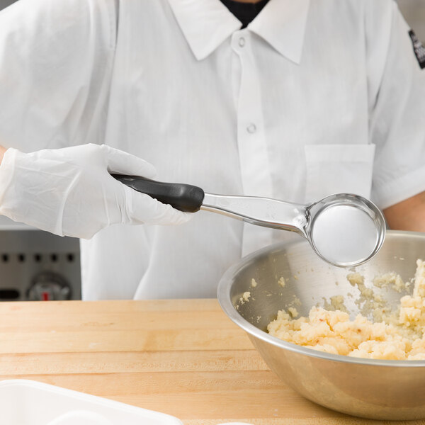 A person in white gloves using a Vollrath black round portion spoon to mix food in a bowl.