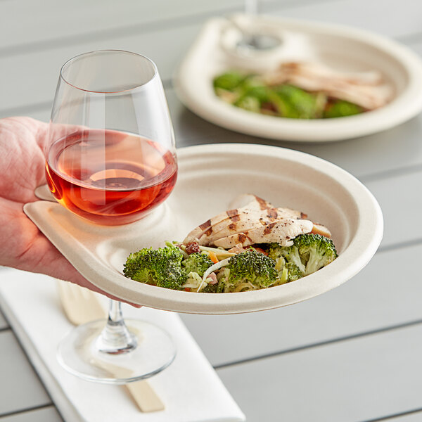A hand holding a World Centric compostable plate with broccoli and chicken and a glass of wine.