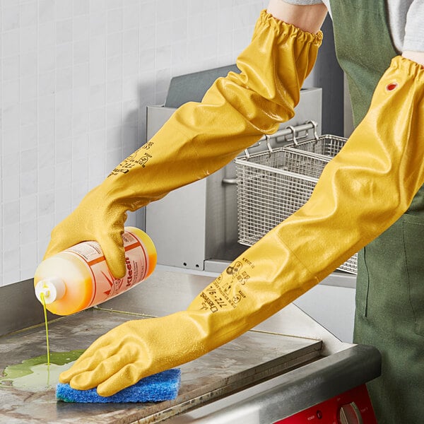 A person wearing Showa yellow knit-lined rubber gloves holding a bottle of cleaning liquid.