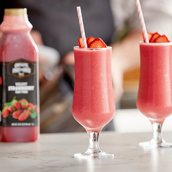 Two glasses of pink strawberry smoothies with straws on a table with a bottle of strawberry syrup.