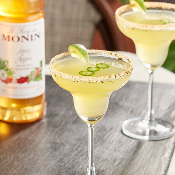 A bottle of Monin Spicy Agave Syrup with two glasses of yellow liquid garnished with limes.