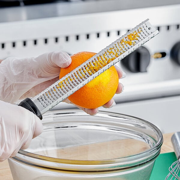 A person using a Microplane zester to peel an orange over a glass bowl.