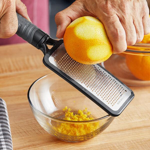 A hand using a Microplane fine grater with a grey handle to zest an orange over a bowl.