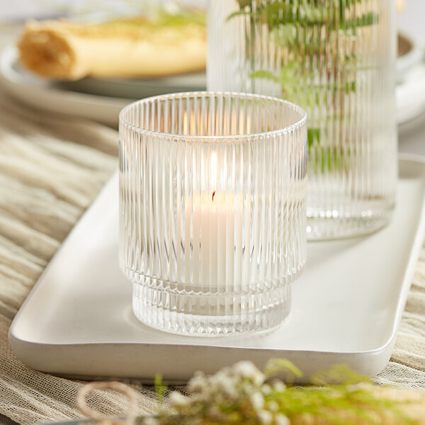 A clear glass Acopa Lore tealight holder with a lit candle inside.