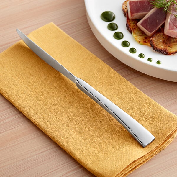 An Acopa stainless steel dinner knife and fork on a table next to a plate of food with a slice of meat.