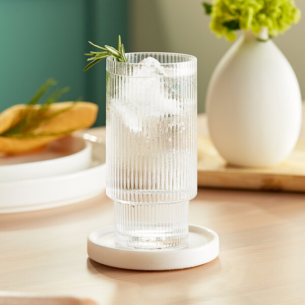 An Acopa Lore highball glass of water with ice and a sprig of rosemary on a coaster.