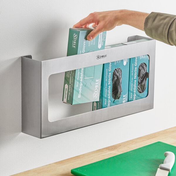 A hand putting a box of Noble Products disposable gloves into a wall-mounted stainless steel box.
