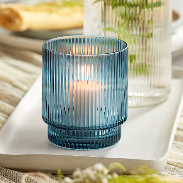 A blue glass Acopa Lore tealight holder with a lit candle on a white tray.