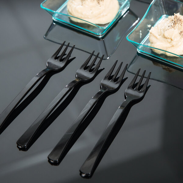 A group of Fineline black plastic tasting forks on a table next to a bowl of hummus.