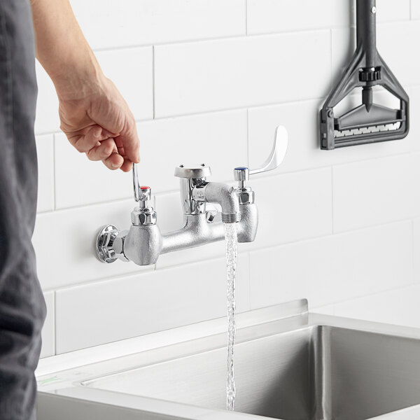 A person using a Waterloo wall-mounted service sink faucet with wrist blade handles to pour water into a sink.