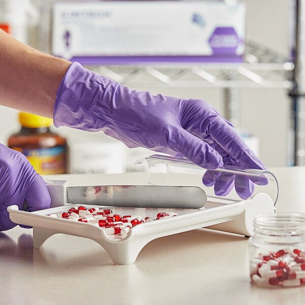 A person in a purple Kimtech nitrile glove cutting red and white pills on a tray.