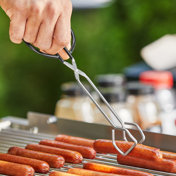A person using Choice 10" Scissor Tongs to hold a hot dog over a grill.