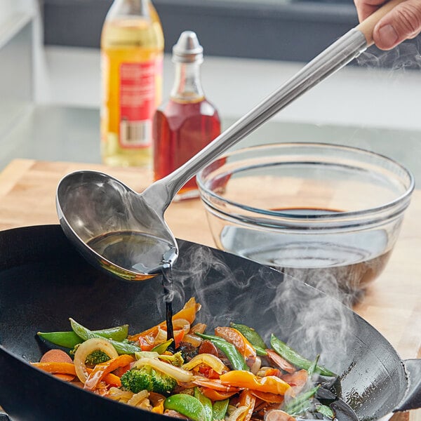 A person using an Emperor's Select small wok ladle to pour liquid into a pan of food.