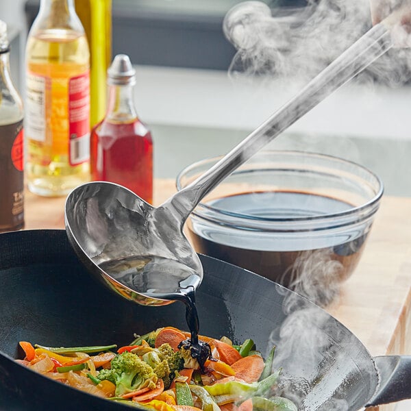 A person using an Emperor's Select large wok ladle to pour liquid into a wok of food.