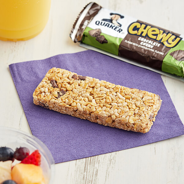 A Quaker Chewy Chocolate Chunk granola bar on a purple napkin next to a bowl of fruit.
