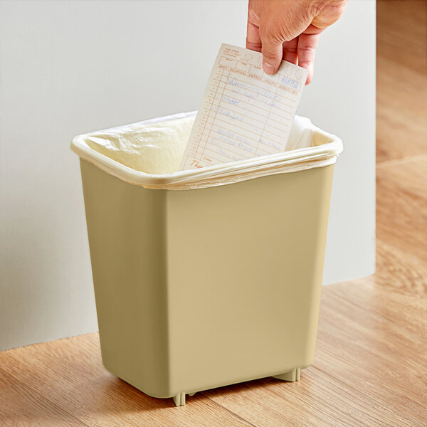 A hand putting a piece of paper in a beige rectangular Lavex trash can.
