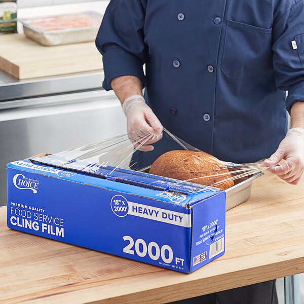 A man in a blue uniform using Choice Heavy-Duty Foodservice Film to wrap a loaf of bread.
