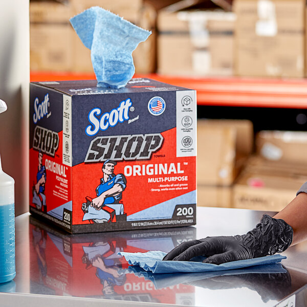 A hand in a black glove using a blue Scott Shop Towel to clean a surface.