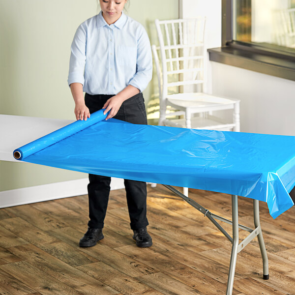 A person rolling a Choice blue plastic table cover sheet onto a table.