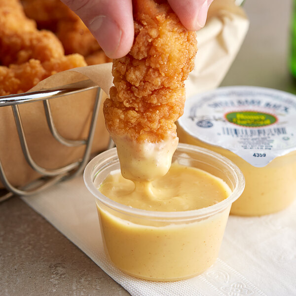 A person dipping a fried chicken nugget into a Sweet Baby Ray's Honey Mustard sauce container.