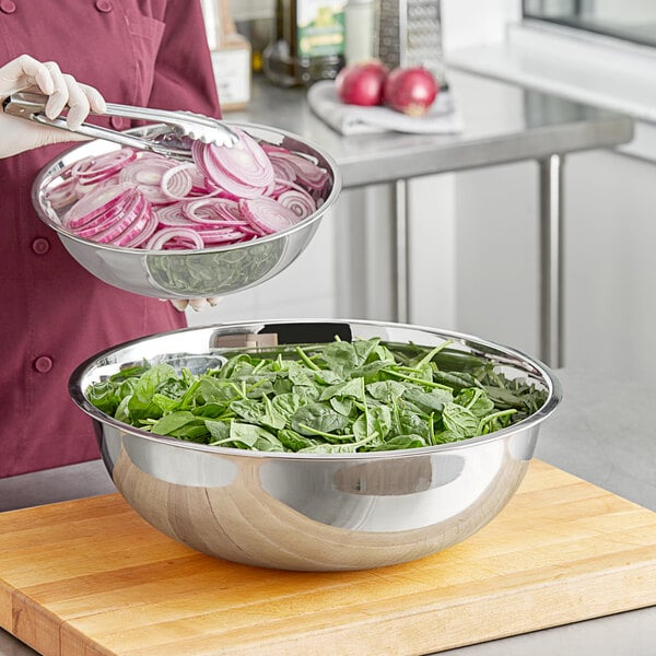 A woman using a Choice 16 Qt. stainless steel mixing bowl to hold sliced red onions and spinach.