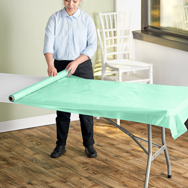 A woman rolling a mint green plastic table cover onto a table.