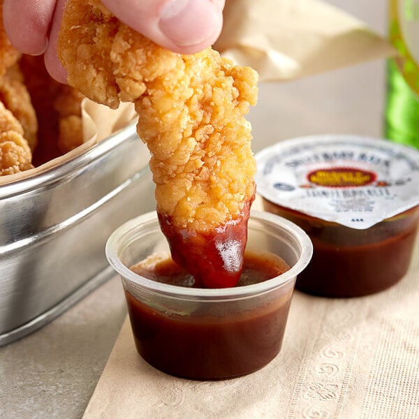 A person dipping a fried chicken strip into a small container of Sweet Baby Ray's BBQ sauce.