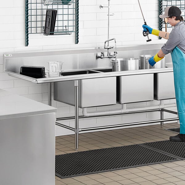 A man in a blue uniform and gloves using a Regency stainless steel 3 compartment sink in a professional kitchen.