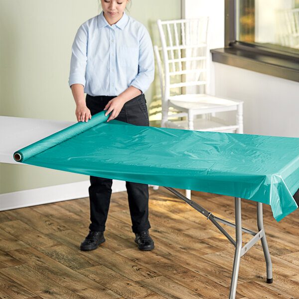 A woman rolling out a teal plastic table cover onto a table outdoors.