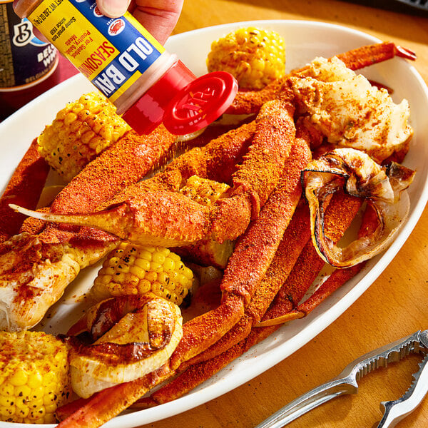 A hand holding a bottle of Old Bay Seasoning over a plate of crab legs and corn.